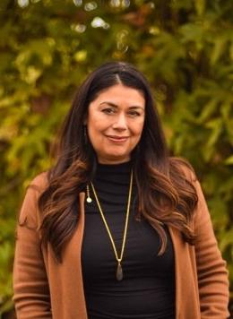 portrait of Yvonne Berumen wearing a black turtleneck and tan cardigan, standing in front of foliage